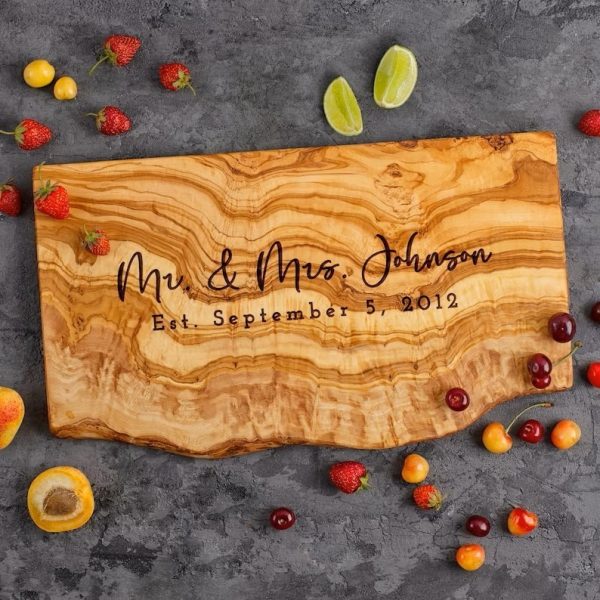 Personalized olive wood live edge cheese board laying on top of a kitchen counter surrounded by pieces of fruit.